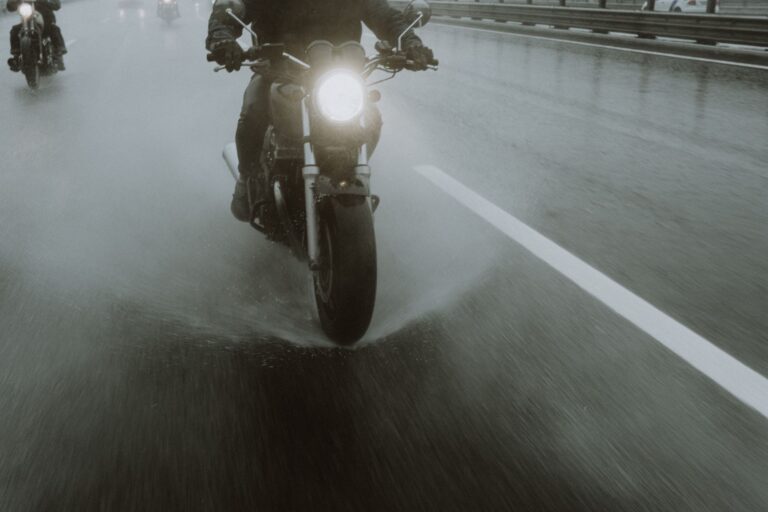 Motociclist echipat cu costum de ploaie care rulează pe un drum ud, cu stropi ridicați de roți